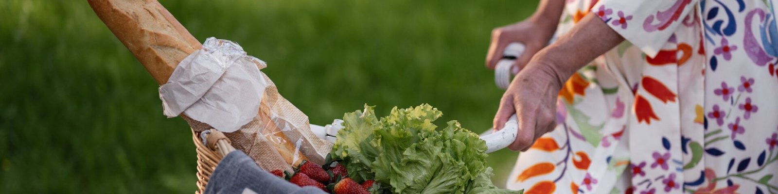A floral-dressed woman with a bicycle basket filled with bread, lettuce, and strawberries outdoors.