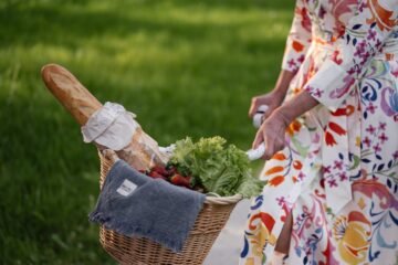 A floral-dressed woman with a bicycle basket filled with bread, lettuce, and strawberries outdoors.