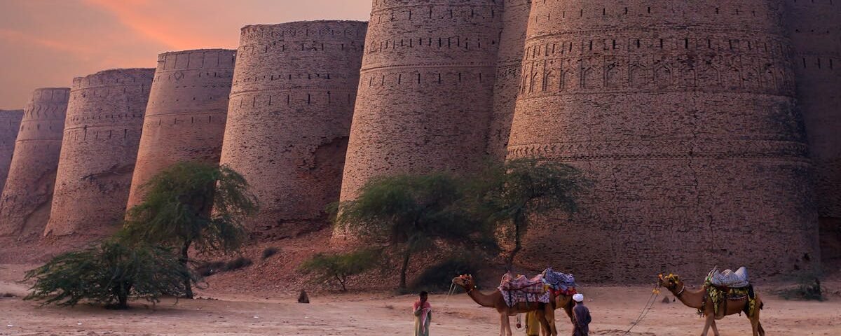 Scenic view of Derawar Fort's towers at sunset in the Punjab desert with camels and people.