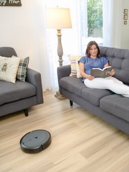 Woman reads on a sofa while a robotic vacuum cleans a brightly lit living room.