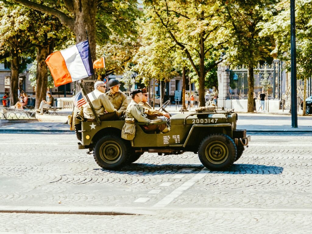 A vintage military jeep with soldiers in a parade, flying French and American flags on a city street.