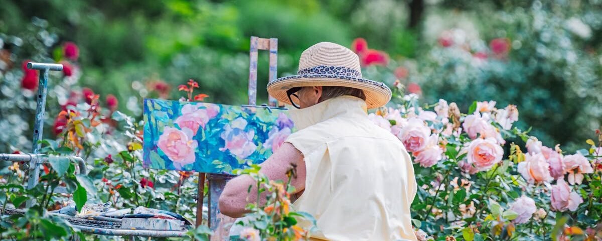 A senior woman painting flowers in a vibrant garden, surrounded by roses on a sunny day.