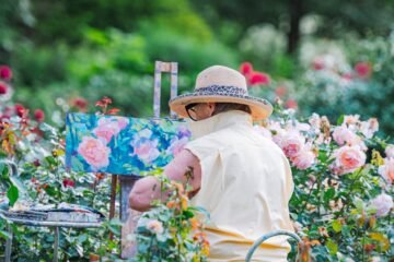 A senior woman painting flowers in a vibrant garden, surrounded by roses on a sunny day.