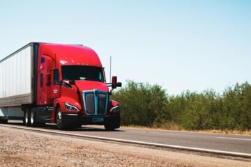 Vibrant red semi-trailer truck driving on an open highway under a clear blue sky.