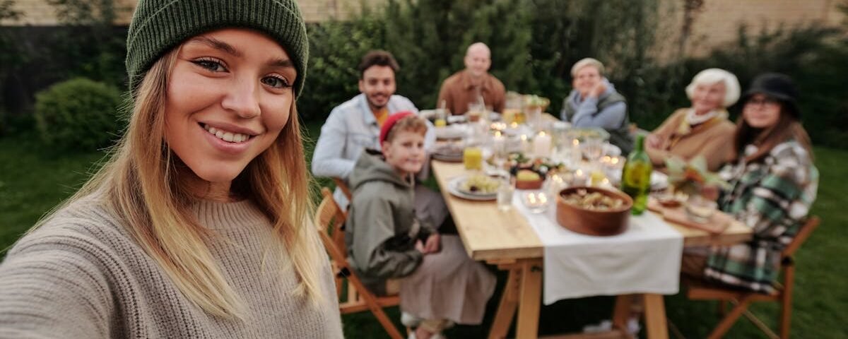 A joyful family dinner outdoors, captured with a smiling woman in the foreground taking a selfie.