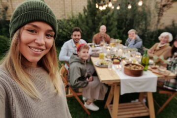 A joyful family dinner outdoors, captured with a smiling woman in the foreground taking a selfie.